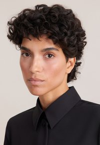 Camicia nera con bottoni e colletto strutturato, dalla texture liscia, indossata da una persona con capelli corti e ricci strettamente arricciati e trucco neutro.