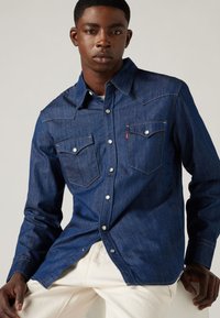 Tumesinise värvusega teksasärk, millel on pikad varrukad, terav krae, kaks rinnataskut, metallist nööpkinnitused ja kontrastne õmblusdetail.