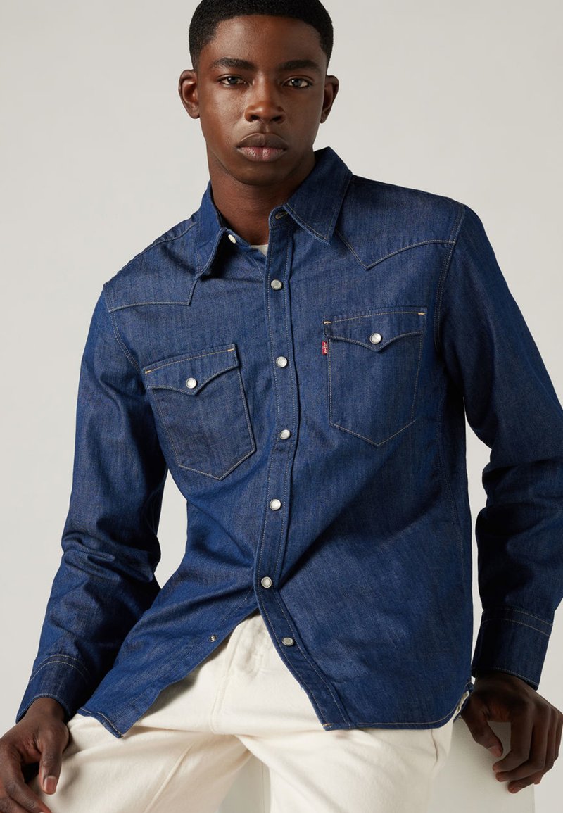 Mörkblå jeansskjorta med lång ärm, spetsig krage, två bröstfickor, metalldragknappar och kontrastsömmar.
