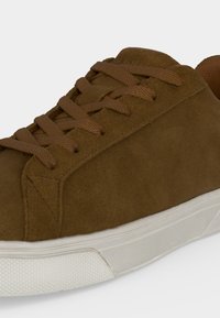Basket en daim marron avec des lacets assortis et une semelle en caoutchouc blanche sur un fond uni.