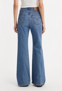 Femme portant un jean bleu taille haute large avec poches arrière et un écusson de marque en cuir marron, debout devant un fond blanc uni.