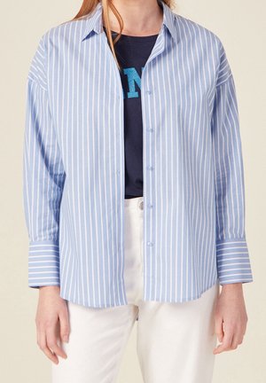 Femme portant une chemise à rayures verticales bleu clair et blanc boutonnée, par-dessus un haut graphique bleu foncé et un pantalon blanc.