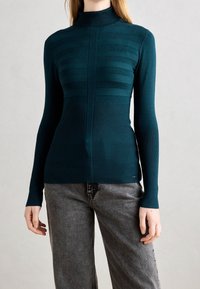 Pull col roulé ajusté couleur sarcelle avec texture côtelée et bandes rayées sur la poitrine, associé à un jean gris à jambes larges.