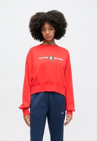 Červený skrátený mikina s grafickým motívom "NIKE SHOX" v bielej farbe, dlhé rukávy a rebrovaný lem. Obliekané s námorníckymi športovými nohavicami.