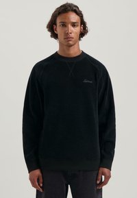 Sudadera de forro polar negra con mangas raglán, puños y dobladillo de canalé. Presenta un sutil bordado de logo en el pecho y una textura suave.