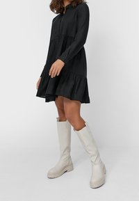 Svart knäppklänning med lagerkant, långa ärmar och en lös passform. Kombinerad med knähöga, beige läderstövlar med en kraftig sula.