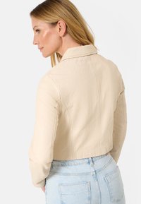 Beige leren cropped jas met een kraag, gestructureerde schouders en een gladde textuur, gecombineerd met lichtblauwe high-waisted spijkerbroek.