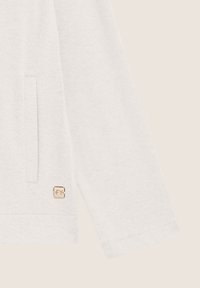 Maglione grigio chiaro con un tessuto testurizzato, dotato di una tasca laterale e di un piccolo logo vicino all'orlo. Maniche lunghe con polsini a costine.