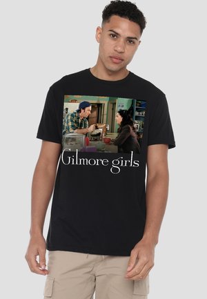 Man wearing black t-shirt with "Gilmore girls" text and café scene of two people talking over coffee printed on front.