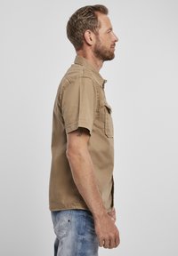 Camicia a maniche corte con bottoni in tessuto marrone chiaro, dotata di taschino sul petto. Presenta un colletto e una vestibilità comoda. Indossata con jeans azzurri.