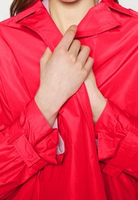 Jasnoczerwone, satynowe okrycie wierzchnie z kołnierzem, marszczonymi mankietami i luźnym krojem. Ręce chwytające odzież, podkreślające jej teksturę.
