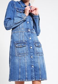 Denim mekko vaaleansinisessä, jossa on napit edessä, pitkät hihat, kaksi rintataskua ja A-linjainen siluetti sauman yksityiskohdilla.