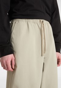Pantalon beige à taille élastique avec cordon de serrage, doté de poches latérales et d'une texture de tissu lisse. Haut à manches longues noir partiellement visible.