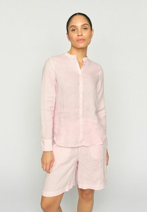 Donna che indossa una camicia rosa chiaro a maniche lunghe con bottoni e pantaloncini abbinati che arrivano al ginocchio, in piedi davanti a uno sfondo chiaro e uniforme.