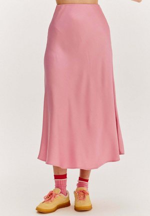 Różowa satynowa spódnica o średniej długości noszona z różowymi i czerwonymi skarpetkami oraz żółtymi trampkami na osobie stojącej na jednolitym tle.