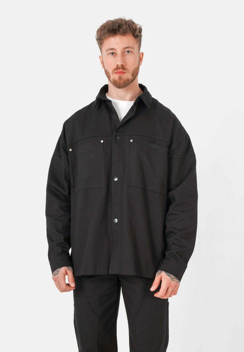 Veste noire en tissu texturé, dotée d'une fermeture à boutons, de deux poches poitrine et de boutons-pression. Coupe décontractée, manches longues.
