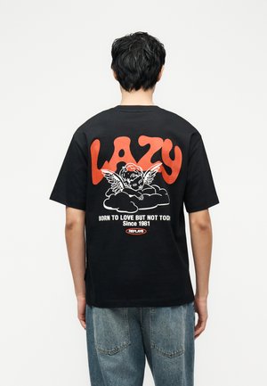 Osoba v čiernom tričku s veľkým oranžovým nápisom „LAZY“, bielym anjelom na oblaku a bielym textom „BORN TO LOVE BUT NOT TODAY.“