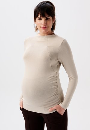 Beige Langarmoberteil mit taillierter Passform, glatter Textur und gerafften Details an den Seiten. Kombiniert mit dunkelbraunen Hosen posiert das Modell mit den Händen in den Taschen.