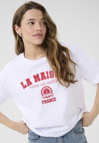 Bijela pamučna majica s crvenim tekstom "LA MAISON VOTRE LIEU PRÉFÉRÉ" i kružnom grafikom s natpisom "FRANCE." Opustitih kroja, kratkih rukava.