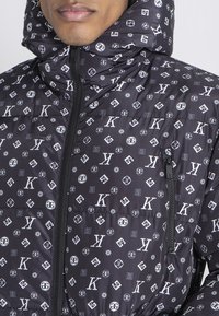 Chaqueta de plumas negra con acabado brillante, que presenta un patrón repetido de logotipos, bolsillos con cremallera y un cuello alto con capucha.