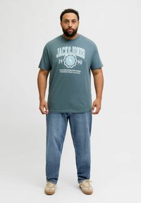 Turkusowa bawełniana koszulka z graficznym nadrukiem, na którym widnieje "JACK&JONES 1990". W połączeniu z jasnoniebieskimi dżinsami i beżowymi trampkami.