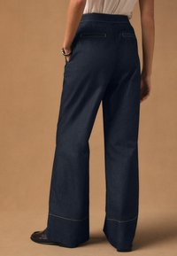 Dunkelblaue, weit geschnittene Jeanshose mit hoher Taille, verziert mit gelben Ziernähten und zwei Gesäßtaschen. Kombiniert mit schwarzen Schuhen.