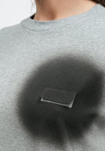 Hägune kaader hallist särgist, millel on väikene kangast tasku rinnal, ümber mille on musta spreivärviga ring.