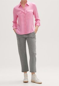 Femme portant une chemise rose à boutons à manches longues, un jean gris droit, et des baskets blanches montantes, debout les mains dans les poches.