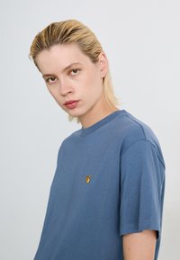 Giovane con capelli biondi bagnati che indossa una maglietta blu Carhartt, che guarda direttamente nella fotocamera contro uno sfondo chiaro e uniforme.
