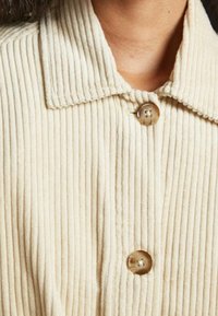 Giacca in corduroy color crema con collo e chiusura a bottoni. Presenta una texture a costine e quattro bottoni lungo la parte anteriore.