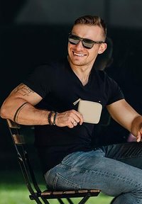 Muž v černém tričku a slunečních brýlích držící světlou peněženku na zip, sedící na dřevěné židli, v modrých džínách a s tetovanými pažemi.