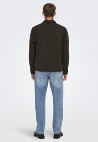 Camisa marrón oscuro, diseño casual con mangas largas y cuello. Llevada con jeans de mezclilla azul claros, que muestran un corte recto y una textura deslavada.