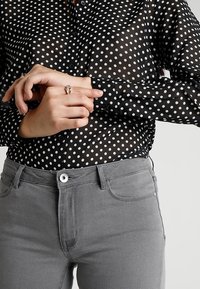 Black sheer blouse with white polka dots, featuring long sleeves and buttoned cuffs, paired with gray fitted jeans.