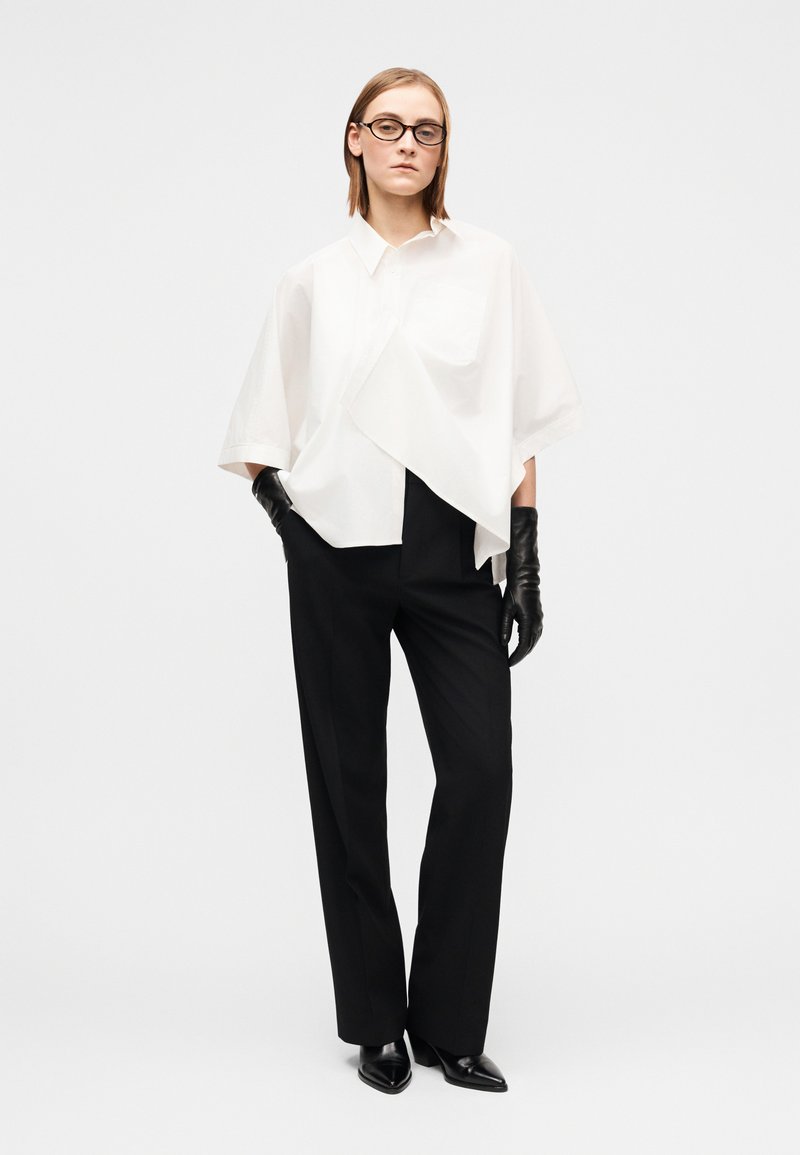 Chemise blanche oversize à manches courtes, pantalon noir sur mesure, gants en cuir noirs et chaussures noires pointues. Design minimaliste.