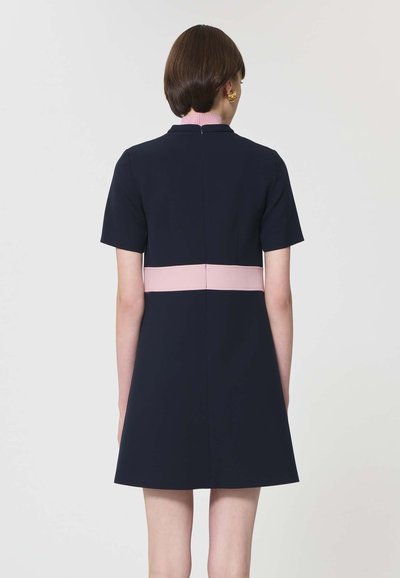 Femme aux cheveux courts bruns portant une robe bleu marine à manches courtes avec une ceinture rose pâle, vue de dos sur un fond uni.