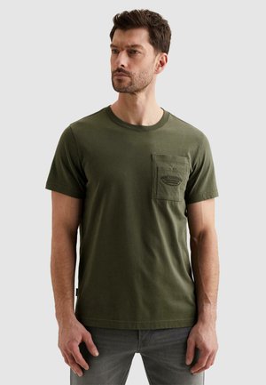 Mężczyzna ubrany w oliwkowo zieloną koszulkę z krótkim rękawem i kieszenią na piersi oraz wyblakłe szare dżinsy, stojący na jasnym, jednolitym tle.