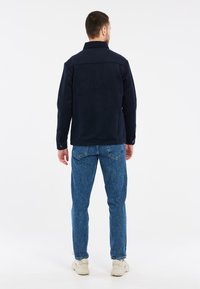 Tamsiai mėlynas polarinis džemperis su apykakle, ilgomis rankovėmis ir sagų rankovėmis, derinamas su šviesiai mėlynomis džinsinėmis kelnėmis ir baltomis sportinėmis bateliais.