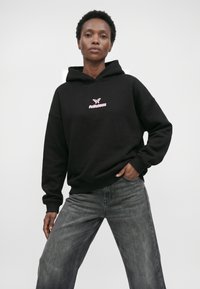 Frau trägt einen schwarzen Hoodie mit einem pinken Schmetterling und dem Schriftzug „felicious“, kombiniert mit locker sitzenden grauen Jeans, steht mit einer Hand in der Tasche.