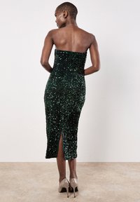 Grünes, mit Pailletten verziertes, trägerloses Kleid mit figurbetonter Silhouette, kniehohem Schlitz hinten und glatter Textur. Kombiniert mit beigen High Heels.