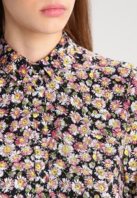 Blusa nera con un motivo floreale, caratterizzata da margherite bianche e rosa con accenti verdi, colletto e dettagli con bottoni sulla parte anteriore.