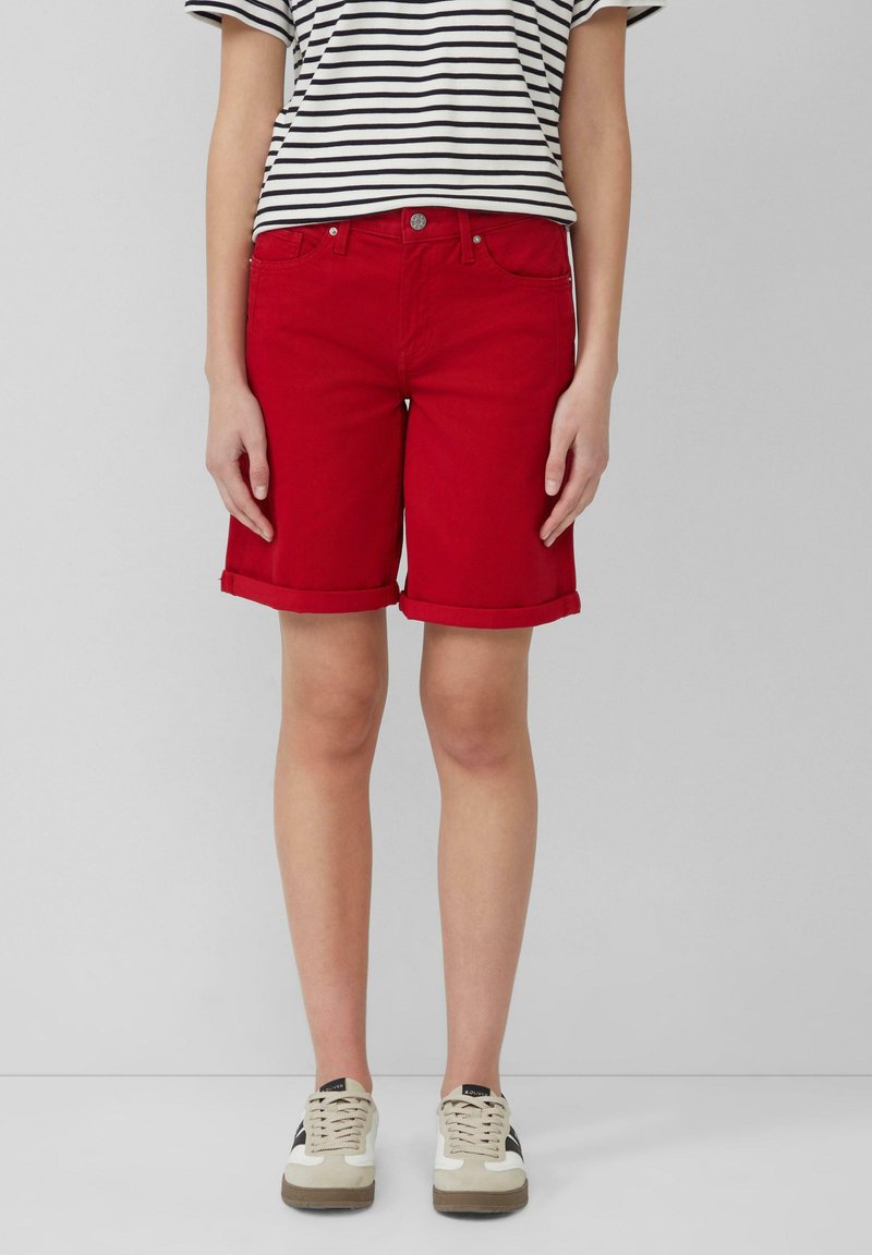 Shorts en denim rouge avec un ourlet roulé, design à cinq poches et fermeture à bouton en métal, associés à un t-shirt rayé noir et blanc et des baskets.