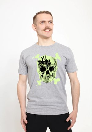 Man met kort haar en snor, gekleed in een grijs T-shirt met een neon groene en zwarte doodskop en gekruiste botten afbeelding, glimlachend en kijkend naar rechts.