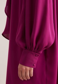Abito magenta a maniche lunghe con polsino arricciato e due bottoni decorativi; tessuto leggero e morbido con una caduta delicata.