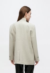 Γυναίκα που βλέπουμε από πίσω, φοράει μπεζ σακάκι με μακριά μανίκια και μαύρο παντελόνι, πάνω από απλό φωτεινό φόντο.