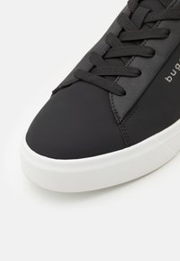 Fekete sneaker sima szintetikus felsőrésszel, fehér gumitalppal és lapos fekete fűzőkkel. Oldalán diszkrét logó található.