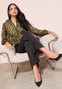 Ženska z dolgimi temnimi lasmi, oblečena v olivno zeleno bluzo z vzorcem, črne široke hlače in črne pete, sedi na teksturiranem fotelju.