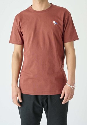 Mężczyzna ubrany w rdzawe krótkie rękawki T-shirt z małą białą i niebieską ptasią haftem na piersi oraz czarne spodnie, stojący na białym tle.