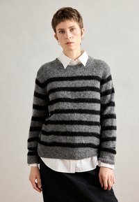 Pulover gri cu dungi negre, realizat dintr-un material texturat, purtat peste o cămașă albă cu guler. Prezintă manșete și tiv cu re configurație.