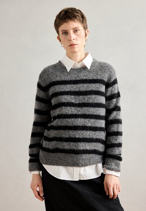 Grijze gestreepte trui met zwarte strepen, gemaakt van een tekstuur materiaal, gedragen over een witte blouse met kraag. Heeft ribbels aan de mouwen en onderrand.
