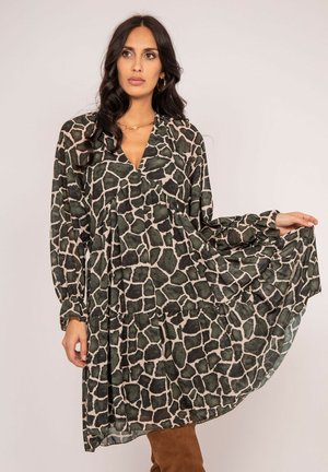 Femme aux longs cheveux bruns portant une robe ample à manches longues avec un imprimé animalier abstrait noir et beige, tenant un côté de la jupe.